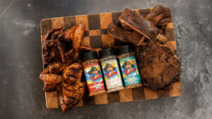 Three of Those Florida Guys BBQ rubs on top of a cutting board surrounded by pieces of meat